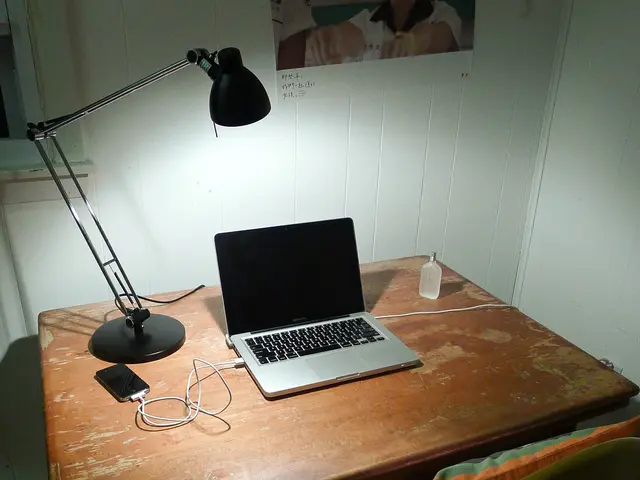 In this image there is a laptop on the table and a light and a mobile phone which is attached to...